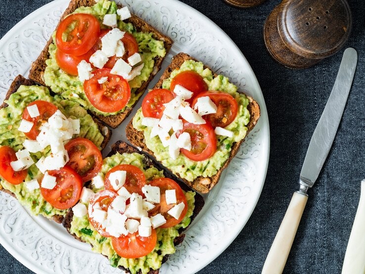 Vier Toasts auf einem weißen Teller belegt mit der Avocado-Basilikum-Creme, Tomate und Mozzarella. Der Untergrund ist eine graue Stoffdecke und neben dem Teller sind Besteck, Salz und Pfeffer. Alles direkt von oben fotografiert.