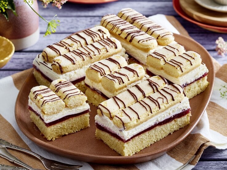 Diese unschlagbaren Schmand-Kirsch-Schnitten dürfen bei deiner nächsten Kuchentafel auf keinen Fall fehlen. Mehrere Schmand-Kirsch-Schnitten auf braunem Teller, im Hintergrund Geschirr und Blumen auf Holzuntergrund.