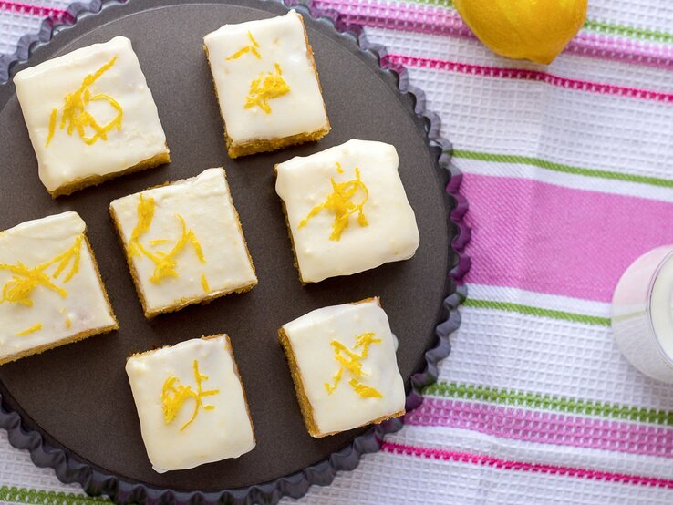 Mehrere Stücke von den Zitronenschnitten mit Limoncello-Guss auf einem dunklen Teller. Darunter eine bunte Tischdecke mit grünen und pinken Streifen.