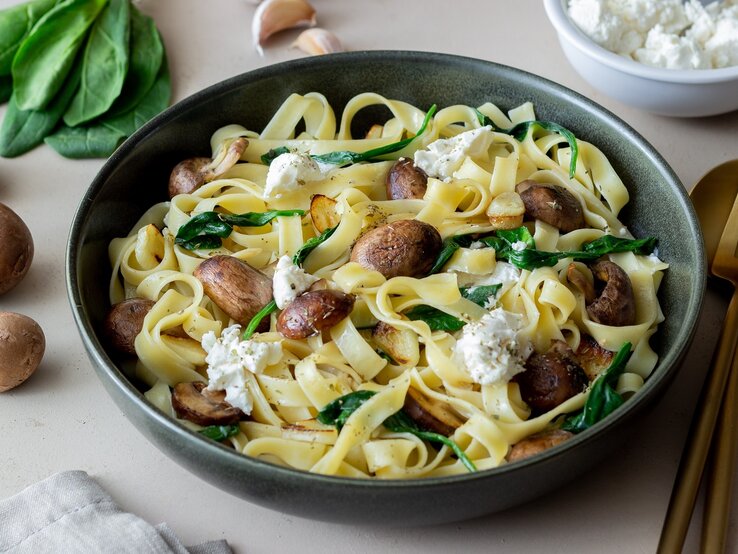 Ein grauer Teller mit dem Pastagericht auf einem hellen Tisch mit Pilzen, Spinat, Feta und Knoblauch.