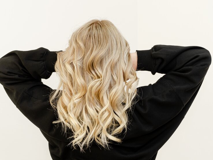 Eine Frau mit blonden Haaren steht mit dem Rücken zur Kamera und fasst in ihre Haare. Sie trägt ein schwarzes Oberteil.