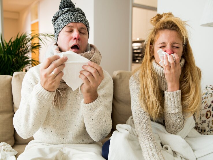 Ein Paar sitzt erkältet auf dem Sofa. Mann und Frau niesen und husten. Die Leute haben eine Erkältung und eine laufende Nase. 