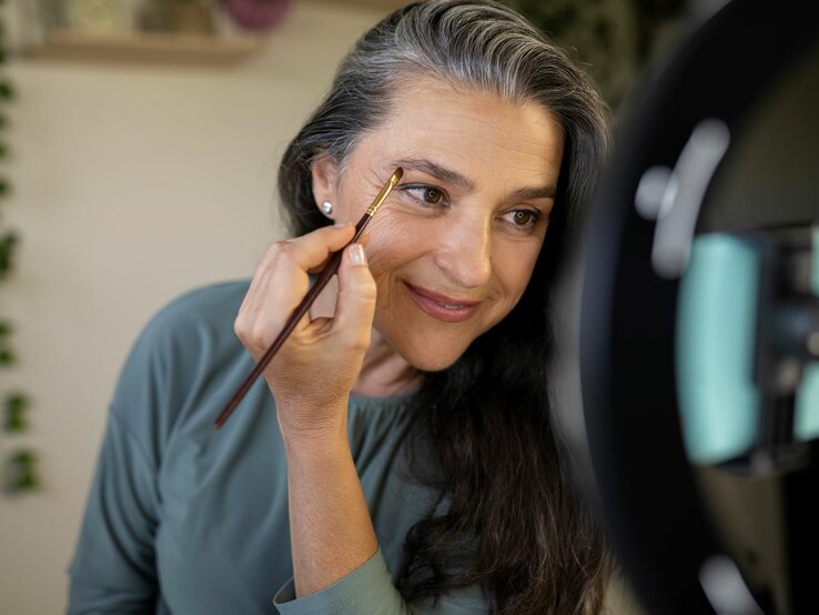 Das Foto zeigt eine Frau mittleren Alters mit graumelierten Haaren, die sich mit einem Make-up-Pinsel die Augenbrauen nachzieht. Sie blickt dabei in einen beleuchteten Kosmetikspiegel. Ihr freundliches Lächeln und der Blickkontakt mit ihrem Spiegelbild vermitteln eine positive und entspannte Stimmung. Sie trägt ein schlichtes, türkisfarbenes Top, das ihre natürliche Schönheit unterstreicht. Die Szenerie deutet auf eine alltägliche Schönheitsroutine hin.