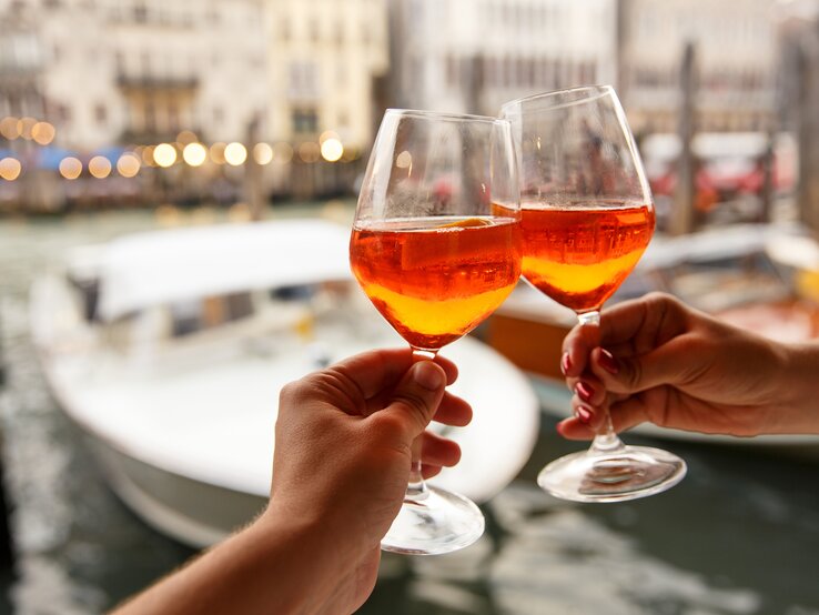 Zwei Hände stoßen mit zwei Gläsern Aperol Spritz an. Im Hintergrund sieht man unscharf die Kulisse eines Hafens. 
