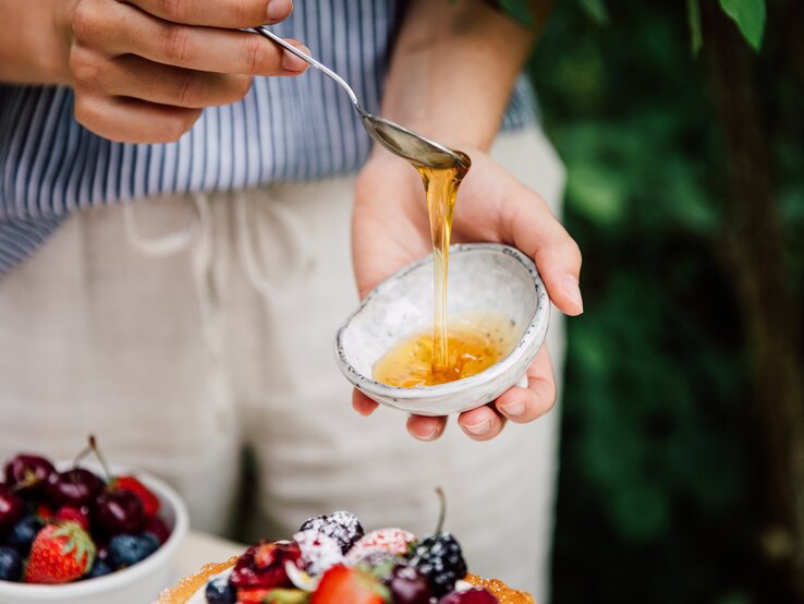 Eine Person gießt mit einem Löffel Reissirup aus einer kleinen Schale. Im Hintergrund unscharf eine Schüssel mit verschiedenen frischen Beeren.