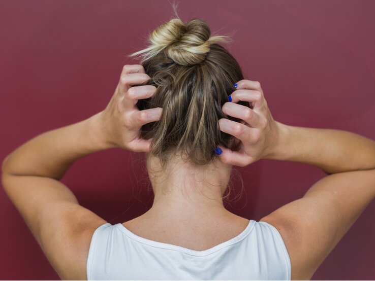 Rückansicht einer Frau, die sich mit beiden Händen an den Kopf fasst. Sie trägt ein weißes Tanktop und hat ihr blondes Haar zu einem lässigen Dutt hochgesteckt. Ihre Fingernägel sind blau lackiert. Der Hintergrund ist einheitlich weinrot, was einen Kontrast zu ihrer hellen Kleidung und Haarfarbe bildet.