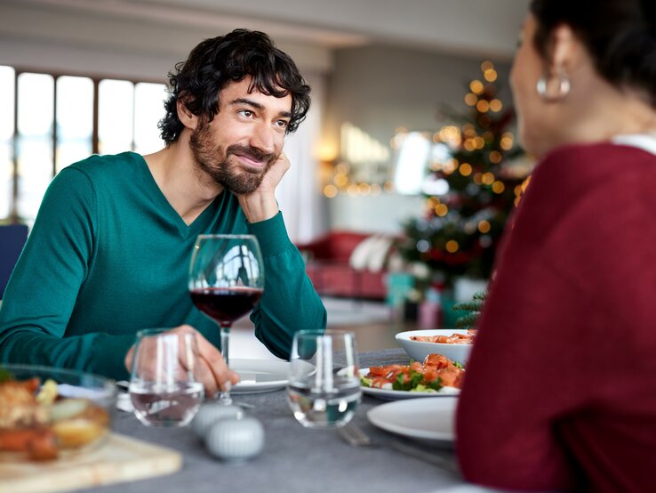 Ein dunkelhaariger Mann in einem grünen Pullover sitzt an einem reichlich gedeckten Tisch und schaut eine dunkelhaarige Frau, die ihm gegenüber sitzt, mit einem liebevollen und zufriedenen Lächeln an. Vor ihm steht ein Glas Rotwein. Der Hintergrund ist unscharf gehalten.