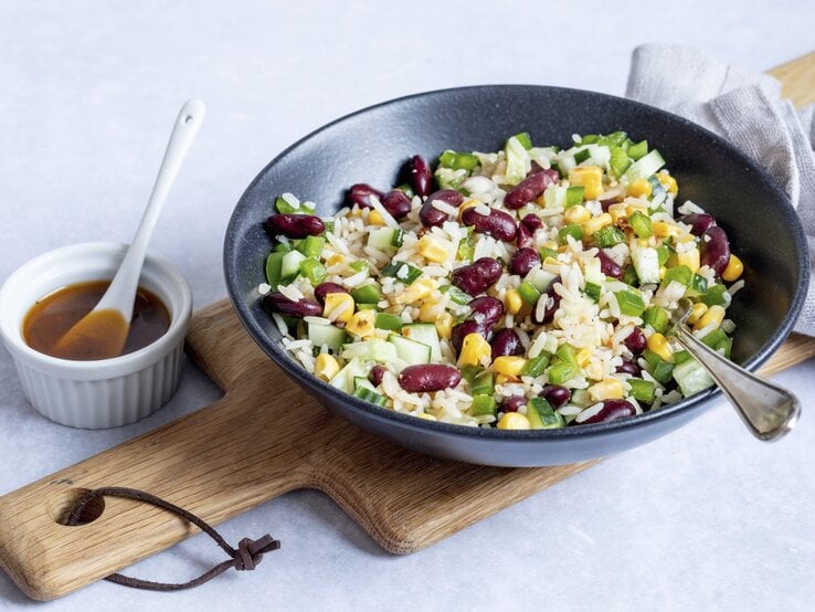 Reissalat mit Mais und Kidneybohnen in einer schwarzen Schüssel. Daneben ein weißes Schälchen mit einem Dressing. 