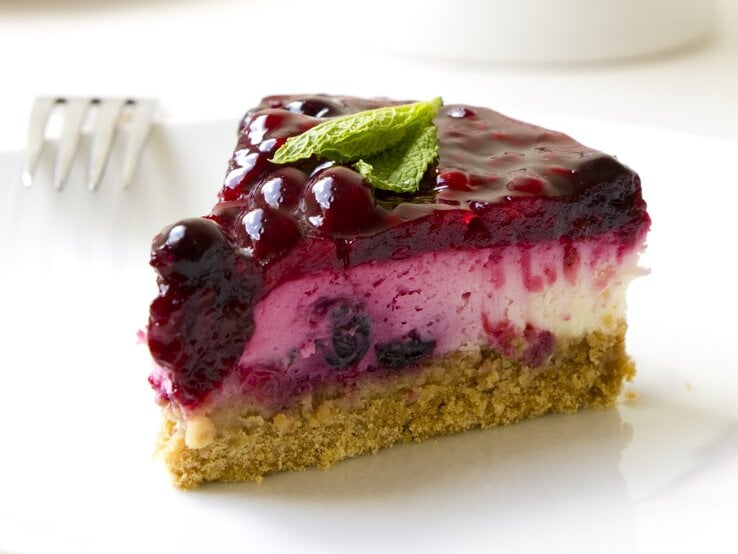 Von unserem Lillet-Käsekuchen reicht niemals nur ein Stück – so köstlich schmeckt er! Angeschnittenes Stück Lillet-Käsekuchen auf weißem Untergrund. Daneben liegt eine Gabel.