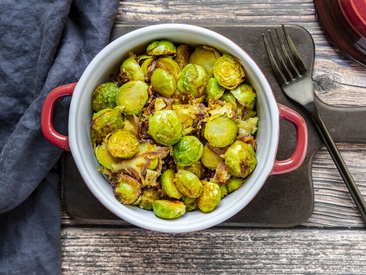 Rosenkohl-Curry In der Draufsicht sieht man einen Topf mit Rosenkohl-Curry auf einem Brett, daneben eine Gabel und ein blaues Geschirrtuch.