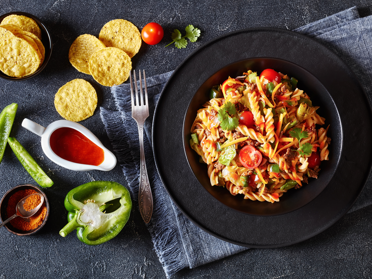 Nudelsalat mit Hackfleisch und Tacos auf schwarzem Teller, angerichtet auf dunklem Untergrund. Daneben liegen verschiedene Zutaten. 