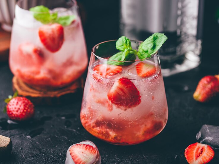 Spritzig-sonnige Leichtigkeit: Ein Erdbeer-Gin-Tonic Erdbeer-Gin-Tonic auf einem dunklen Untergrund. Daneben liegen verschiedene Zutaten.
