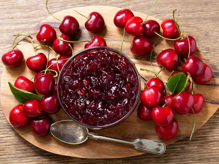 Pack den Sommer ins Glas: eine selbstgemachte Kirschmarmelade. Auf einem Holzbrett steht ein Glas Kirschmarmelade. Drum herum liegen Kirschen.