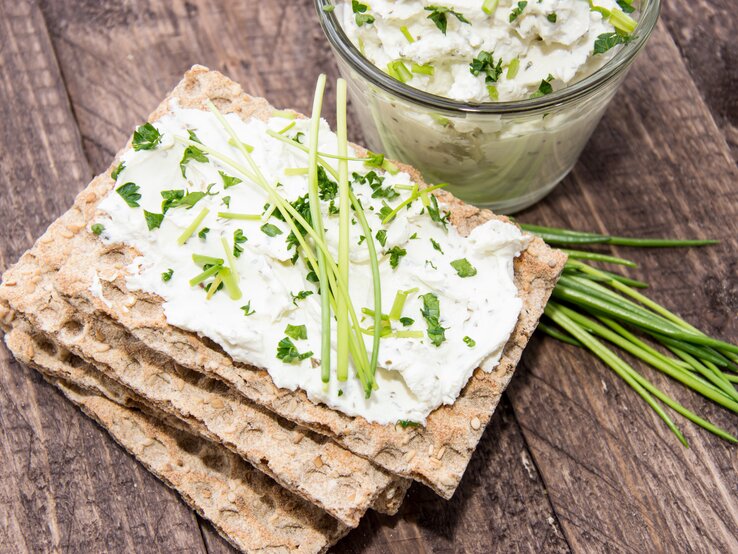 Draufsicht. Vier Scheiben Knäckebrot liegen auf einem Untergrund aus Holz. Auf der obersten Scheibe ist ein heller Aufstrich aus Frischkäse mit frischen Kräutern, wie etwa Schnittlauch und Petersilie.