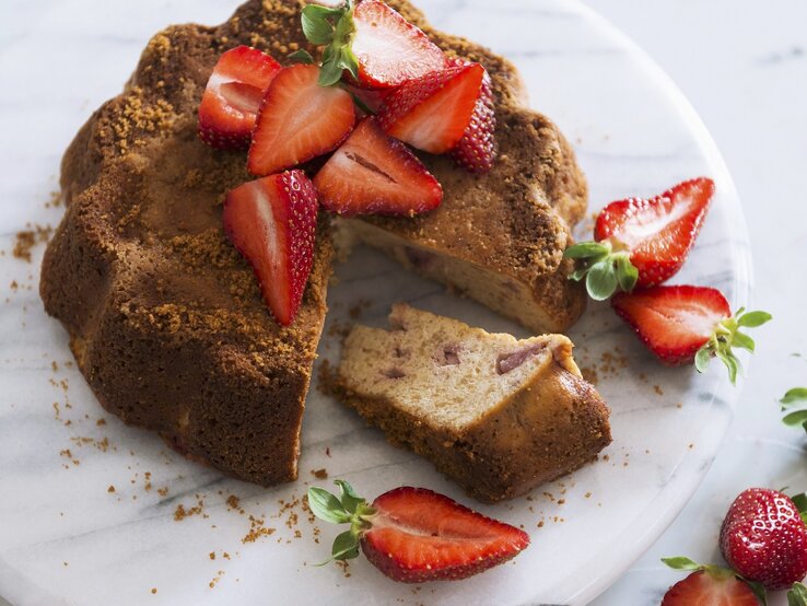 Ein angeschnittener Sauerrahmkuchen mit Erdbeeren auf einem hellen Marmorteller und weißem Untergrund.