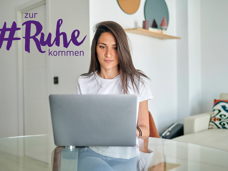 Eine junge Frau sitzt konzentriert an einem gläsernen Tisch und arbeitet an ihrem Laptop. Sie trägt ein weißes T-Shirt und hat lange dunkle Haare. Im Hintergrund ist ein modern eingerichtetes Zimmer mit hellen Wänden und zeitgenössischer Dekoration zu sehen. Auf dem Laptop ist der Schriftzug "#zur Ruhe kommen" zu sehen.