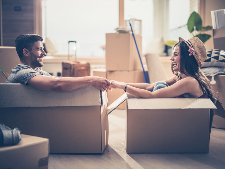 Junger Mann und junge Frau sitzen lachend jeweils in einem Umzugskarton in einer Wohnung