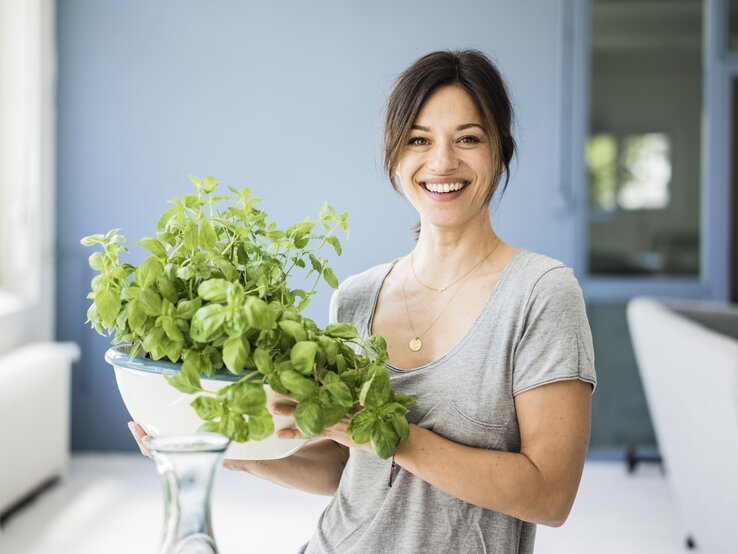 Eine fröhliche Frau hält eine Schale mit üppigem grünem Basilikum. Sie trägt ein graues T-Shirt und steht in einer hellen, freundlich eingerichteten Küche mit blauen Wänden. Ihr Lächeln und die frischen Kräuter in ihren Händen vermitteln ein Gefühl von Gesundheit und Freude am Kochen.