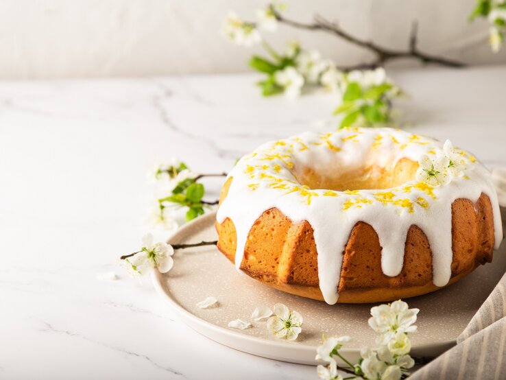 Ein Aperol-Gugelhupf auf einem runden Teller, der Kuchen mit Zuckerguss und Zitronenabrieb dekoriert, im Hintergrund Zweige und Blüten.