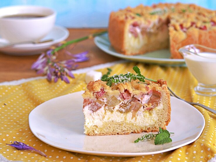 Ein Stück vom Rhabarberkuchen auf einem weißen Teller und einer gelb-blauen Tischdecke. Dahinter eine Tasse Kaffee und der angeschnittene Kuchen. 