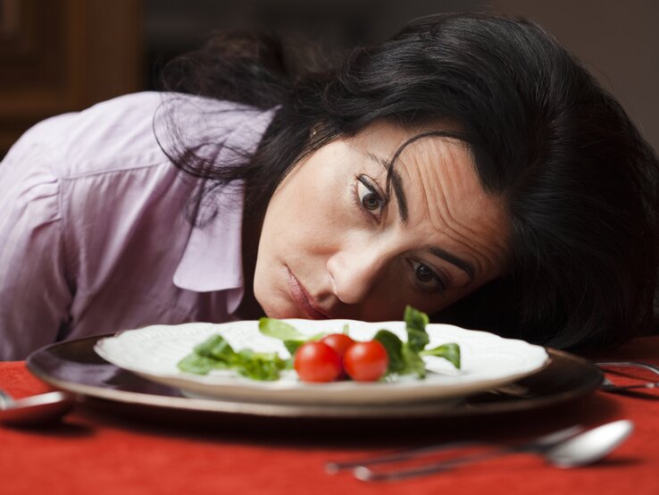 Eine Frau mittleren Alters mit dunklen Haaren und einem nachdenklichen Blick liegt mit ihrem Kopf auf einem Tisch und schaut auf einen Teller mit einer sehr kleinen Menge Essen, bestehend aus einigen Blättern Salat und zwei Kirschtomaten. Ihr Gesichtsausdruck könnte Enttäuschung, Unzufriedenheit oder Desinteresse an der spärlichen Mahlzeit ausdrücken. Sie trägt eine hellviolette Bluse, und das Ambiente wirkt häuslich. Der rote Tischläufer unter dem Teller verleiht dem Bild eine warme Atmosphäre.