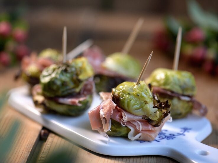Herzhafter Snack: Rosenkohl-Spießchen mit Salami und Prosciutto Rosenkohl-Spießchen mit Salami und Prosciutto auf einem weißen Brett mit unscharfem Hintergrund.