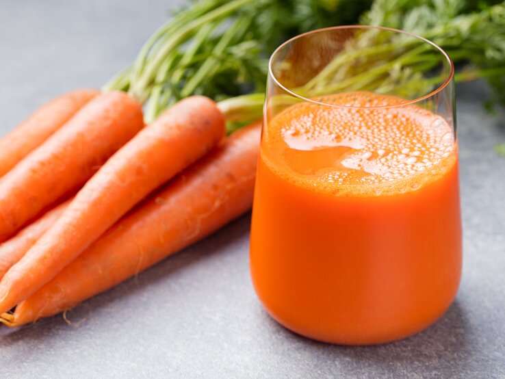 Ein Bild von einem Glas voll frisch gepresstem Karottensaft auf einer grauen Unterlage. Hinter dem Glas befindet sich ein Bündel ganzer, ungeschälter Karotten mit grünen Stielen, die leicht unscharf sind. Der Saft hat eine leuchtend orange Farbe, und es sind Blasen an der Oberfläche zu sehen, was darauf hindeutet, dass er frisch ist. Das Bild konzentriert sich auf das Glas im Vordergrund, während die Karotten im Hintergrund eine Idee der Herkunft des Saftes geben.