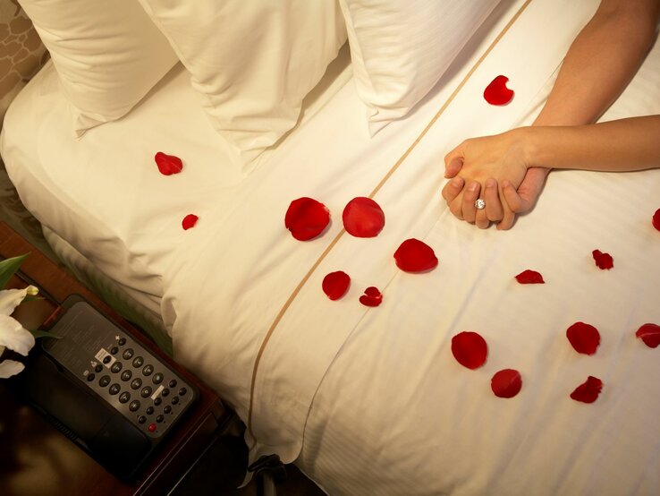 ein weißes Bett mit Rosenblüten bestreut, die verschränkten Hände eines Paares sind zu sehen