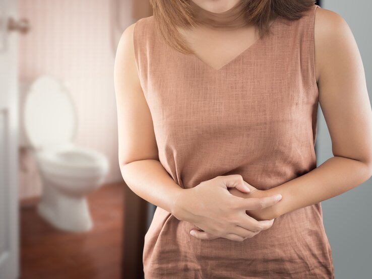 Frau hat eine Blasenentzündung und hält sich den Bauch. Im Hintergrund sieht man das Badezimmer und die Toilette. 