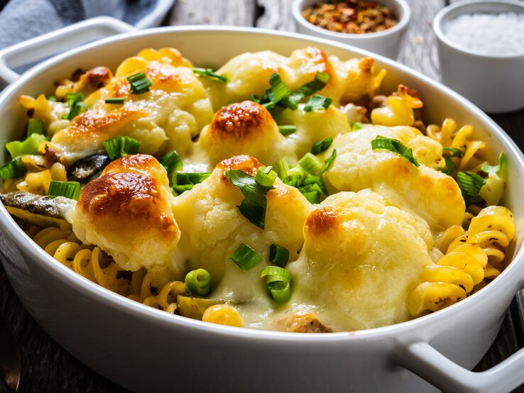 Schräg oben: ein überbackenes Blumenkohl-Gratin mit Hackfleisch und Käse in einer weißen Auflaufform steht auf einem Holztisch.