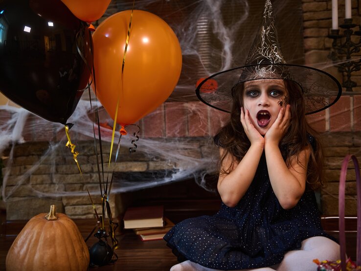 Bezauberndes kleines Mädchen mit kaukasischer Herkunft, gekleidet in eine Hexen- und Zaubererhut, sitzt neben orangefarbenen und schwarzen Luftballons und einem Kürbis, vor einem mit Spinnweben bedeckten Kamin. Halloween-Party. Gotische Festlichkeit. 31. Oktober.