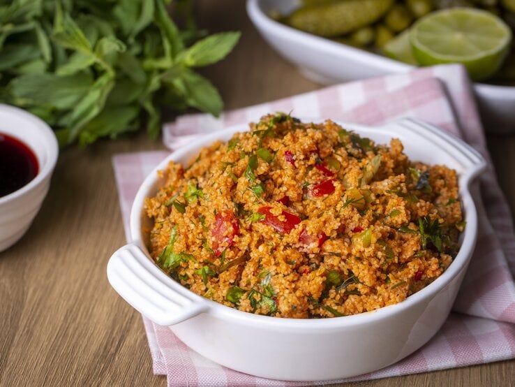 Frontalsicht: Auf einem Holztisch steht eine Schüssel Kisir, einen türkischen Bulgur-Salat. Dahinter liegen frische Minze, Zitronen und eine Schale mit einer roten Flüssigeit.