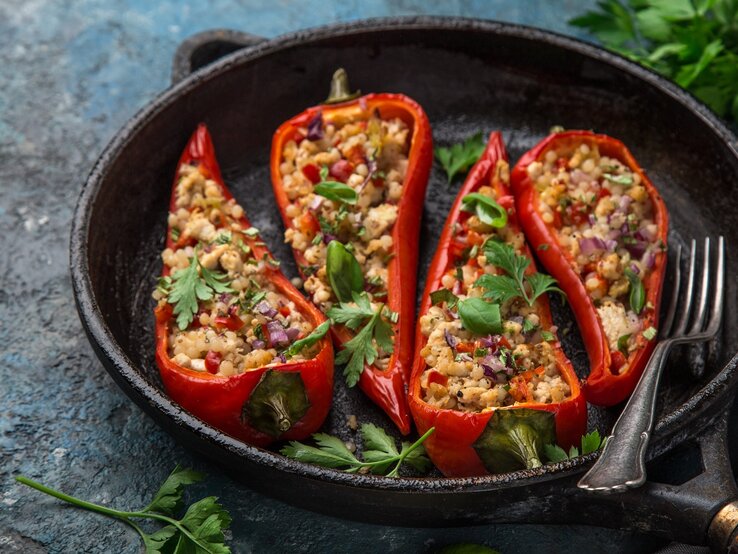 Gefuellte-Paprika-Perlen-Couscous Eine gusseiserne Pfanne mit den vier gefüllten udn geschmorten Paprikaschoten auf einem dunkelgrauen Tisch mit Petersilie und einem grauen Tuch.