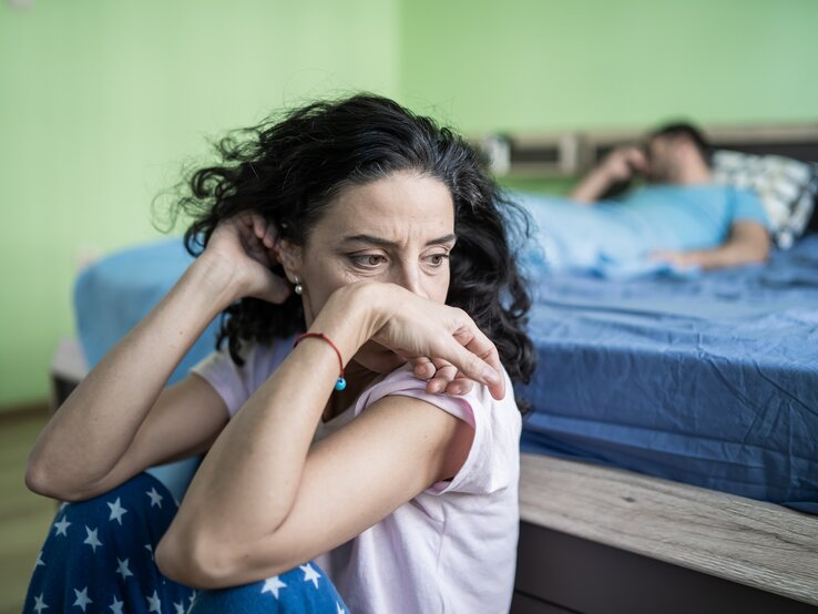 Eine Frau sitzt resigniert vor dem Ehebett, im Bett liegt ein Mann und döst oder denkt nach | © iStock.com/valentinrussanov