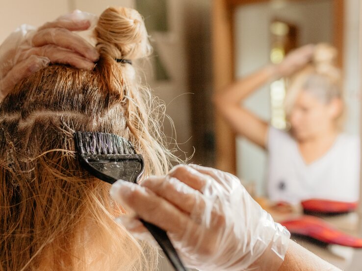 Frau färbt sich ihre Haare vor dem Spiegel mithilfe eines Pinsels, sie trägt Einmalhandschuhe