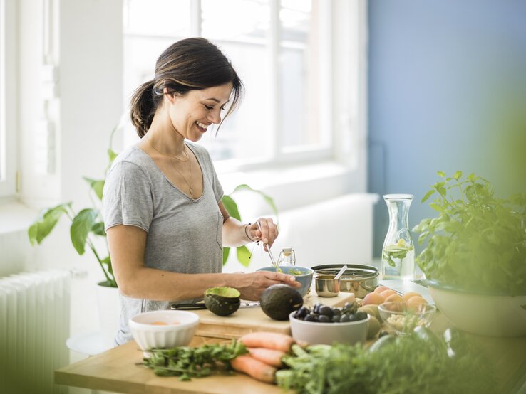 Eine Frau mit dunklem Haar und einem grauen Shirt bereitet in einer hellen Küche Essen vor. Sie lächelt, während sie an einem Holztisch steht, auf dem verschiedene frische Zutaten zu sehen sind: Avocados, Karotten, Oliven und diverse Kräuter. Im Hintergrund sind helle Fenster und ein lebendiges Interieur zu erkennen. Die Szene strahlt Wohlbefinden und die Freude am Kochen mit frischen Produkten aus.