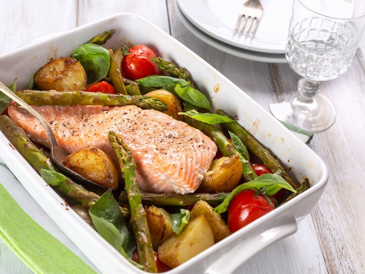 Eine weiße Auflaufform mit dem Ofenlachs auf einem weißen Holztisch. Daneben ein schickes Glas und zwei weiße Teller.