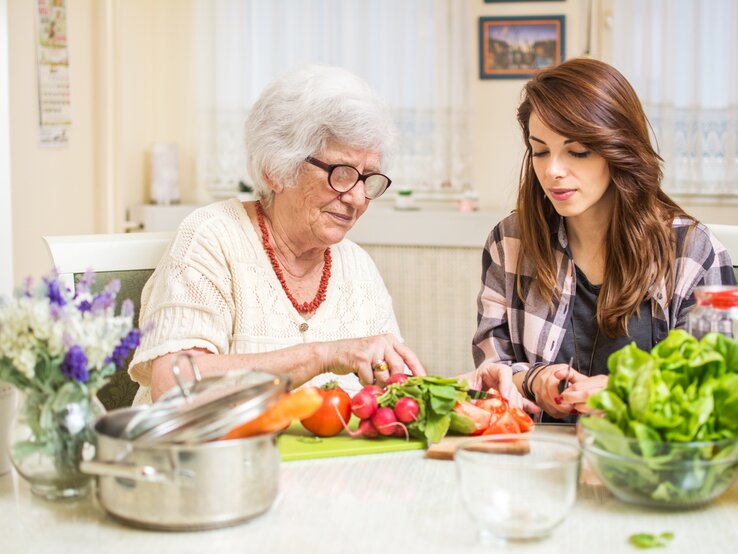 Eine ältere Dame mit weißen Haaren und Brille bereitet gemeinsam mit einer jüngeren Frau mit braunen Haaren und kariertem Hemd frisches Gemüse in einer heimeligen Küche zu. Auf dem Tisch sind eine Schüssel mit grünem Salat, frische Tomaten, Radieschen und weitere Küchenutensilien zu sehen. Hinter ihnen befindet sich ein Fenster, das den Raum mit natürlichem Licht erfüllt, und auf dem Tisch steht ein kleines Bouquet aus lila und weißen Blumen. Das Bild strahlt eine warme, familiäre Atmosphäre aus und zeigt Generationen, die gemeinsam kochen.