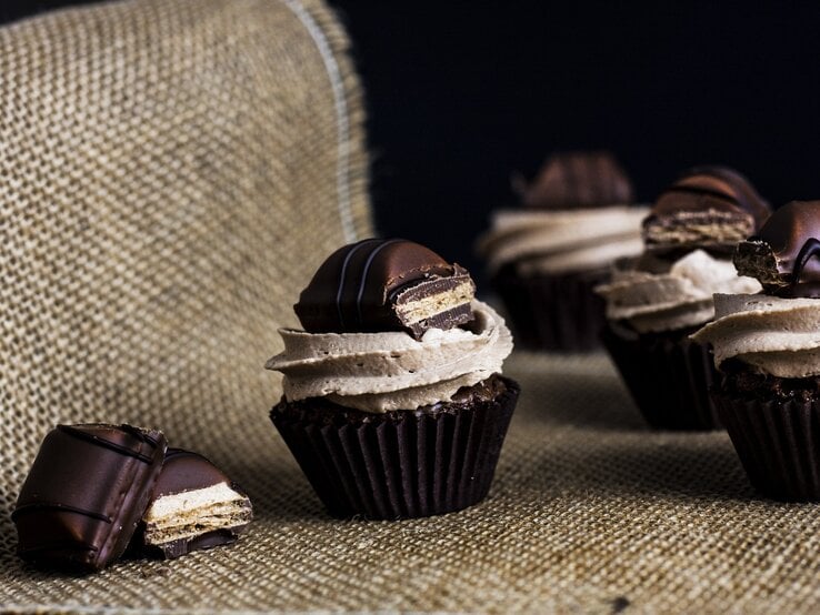 Kinder-Bueno Cupcakes mit Milchmädchencreme auf Jutebeutel vor schwarzem Hintergrund. Daneben Schokoriegel-Stücke. Frontalansicht. 
