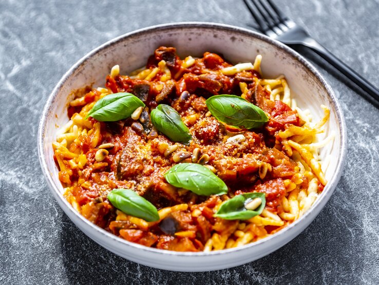 Ein Teller Pasta mit Frischkäsesoße, Basilikum und Pinienkernen, daneben liegt eine Gabel.