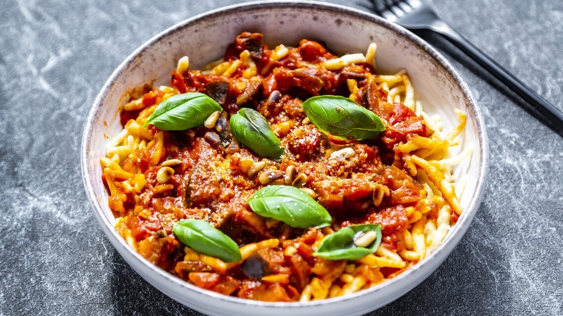 Ein Teller Pasta mit Frischkäsesoße, Basilikum und Pinienkernen, daneben liegt eine Gabel.