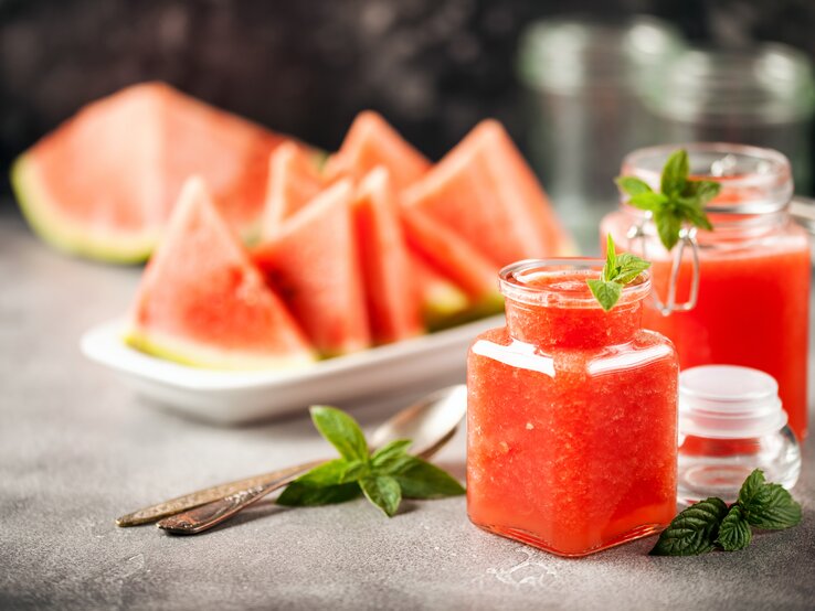 Zwei Gläser mit Wassermelonen-Minz-Gelee stehen auf einem grauen Untergrund. Daneben liegen Minz-Blätter und Löffel. Eine Schale mit Melonenstücken liegt im Hintergrund.