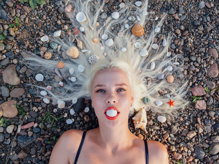 Eine blonde Frau liegt mit dem Rücken auf einem Kieselstrand, umgeben von verschiedenen Meeresmuscheln, die in ihrem ausgebreiteten Haar arrangiert sind, während sie eine Muschelschale zwischen ihren Lippen hält, die wie ein Mund aussieht.