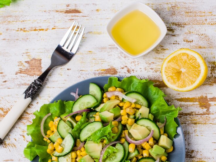 Schale mit Mango-Dressing neben einem Teller Salat mit Gurke und Mais auf hölzernem Untergrund. 