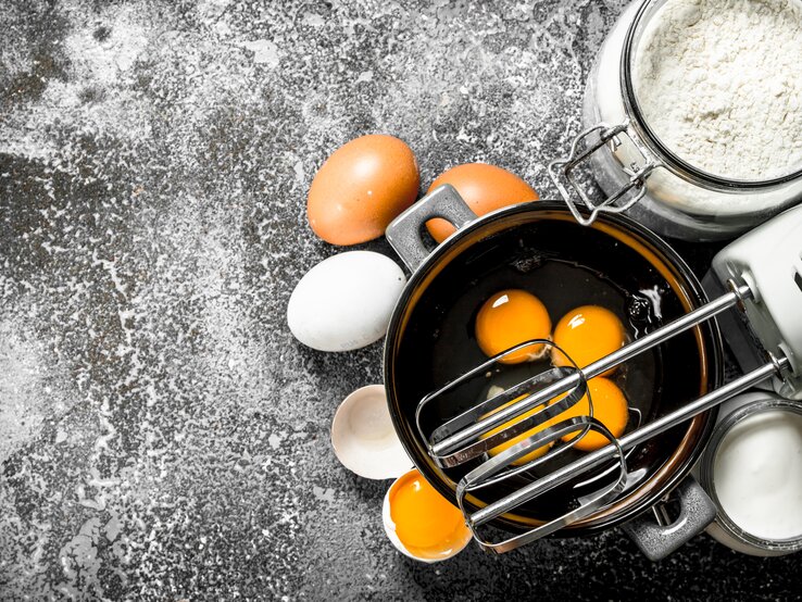 Draufblick: Auf einer dunklen bemehlten Arbeitsfläche steht eine Schüssel mit Eiern. Daneben sind ein Handmixer, Eier, Mehl, Sahne und Eierschalen zu sehen.