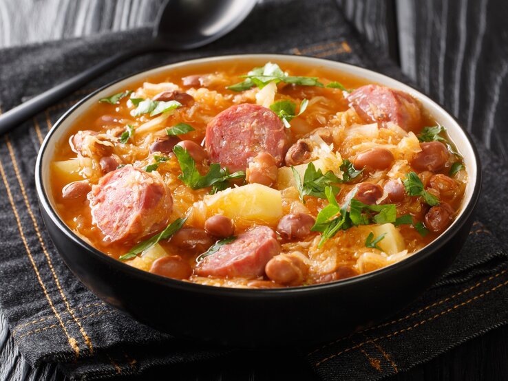 Sauerkraut-Eintopf mit Steckrüben und Räucherwurst in Suppenschüssel auf grauem Tuch. Daneben Suppenlöffel. Draufsicht.