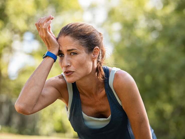 Eine erschöpfte sportliche Frau mittleren Alters mit einem Fitness-Tracker am Handgelenk wischt sich Schweiß von der Stirn, während sie sich in einer natürlichen grünen Umgebung erholt. Sie trägt ein ärmelloses Top und sieht aus, als hätte sie gerade ein intensives Training absolviert. Der Ausdruck auf ihrem Gesicht spiegelt Anstrengung und Konzentration wider.