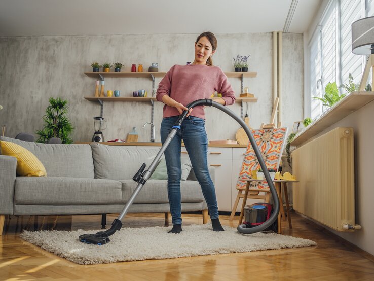 Eine Frau steht mit einem Staubsauger auf einem Teppich in einem gemütlichen Wohnzimmer und hält sich dabei den Rücken, als hätte sie Schmerzen. Sie trägt ein rosa Oberteil und Jeans und hat einen schmerzverzerrten Gesichtsausdruck, der auf Beschwerden beim Hausputz hinweisen könnte. Das Zimmer ist hell und einladend, mit einer Couch, Pflanzen, und einer modernen Einrichtung.