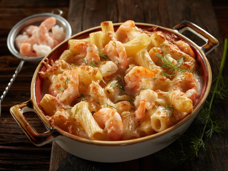 Frontalsicht: Auf einem dunklen Tisch steht eine dunkle Keramik-Auflaufform mit überbackener Pasta mit Dill-Schmand-Garnelen. Daneben liegt ein kleines Sieb mit rohen Garnelen.