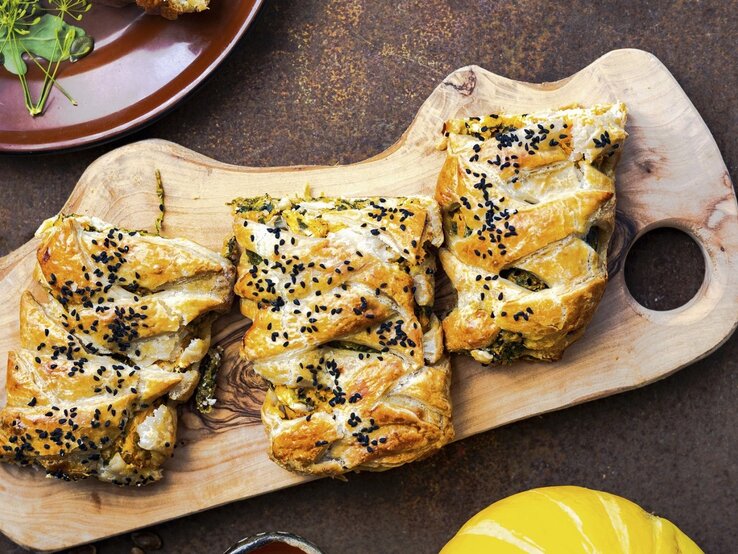 Blaetterteiggebaeck-Kuerbis-Spinat-Feta Auf einem Schneidebrett liegt Blätterteiggebäck gefüllt mit Kürbis, Spinat und Feta. Es ist mit Sesam bestreut.